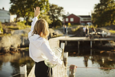 Rear view of young woman waving to friends standing on jetty at harbor