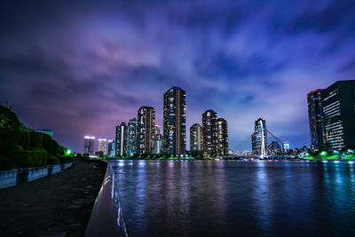 Illuminated modern buildings by sea against sky at night