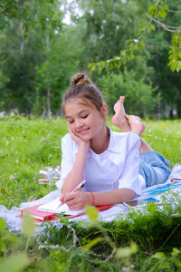 A young schoolgirl lies on the grass in the park with a notebook and a pen.