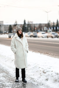 Girl in a fur coat walks along the road traffic of cars. the girl has a knitted hat on her head