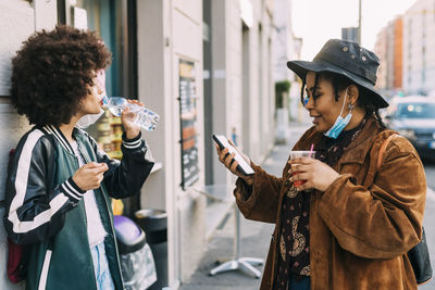 Young woman drinking water by friend using mobile phone on footpath