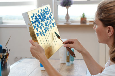 Woman artist painting picture at home with acrylic paints and spatula over the window. adult hobby