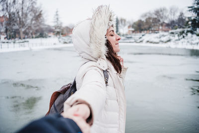 Happy caucasian woman holding hand of boyfriend, standing in front of frozen lake during winter