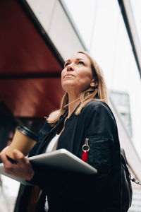 Low angle view of mature businesswoman walking with laptop and coffee cup in city