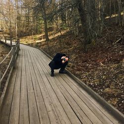 Man photographing through camera while crouching on boardwalk at forest