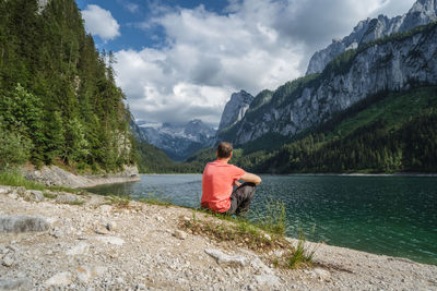 Rear view of man sitting on rock by lake against mountains