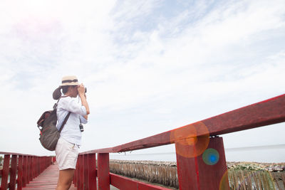 Side of woman photographing on boardwalk against sky