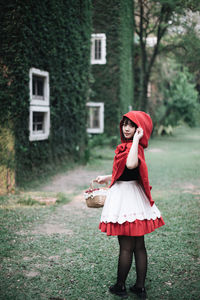 Full length of woman in little red riding hood costume holding basket while standing against building