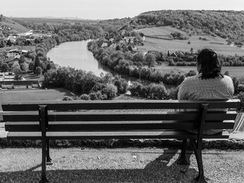 Rear view of woman sitting on bench against river and landscape