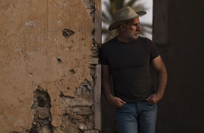 Portrait of adult man in cowboy hat and jeans against abandoned building during sunset