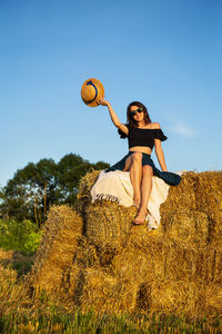 A young woman looks at the setting sun and holds a straw hat, sits on a pile of straw bales.