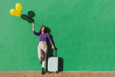 Modern fashionable carefree female holding bunch of yellow balloons and suitcase while standing against green wall in sunny day