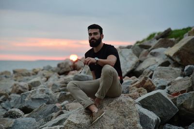 Young man sitting on rock at beach against sky during sunset