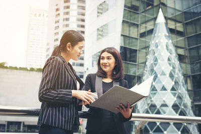 Smiling businesswoman with folder looking at colleague while standing against buildings 