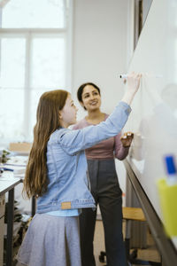 Side view of schoolgirl writing on whiteboard with felt tip pen by teacher in classroom