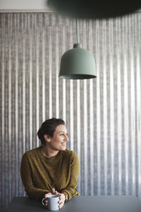 Smiling businesswoman sitting with coffee cup at desk against corrugated iron wall