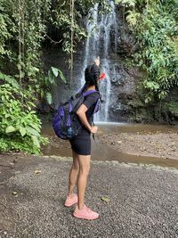 Full length side view of woman standing by waterfall in forest