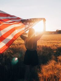 Rear view of woman standing on field against sky during sunset holding us flag 