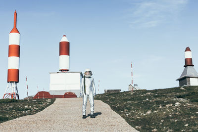 Full body man in spacesuit standing on rocky ground against striped rocket shaped antennas on sunny day