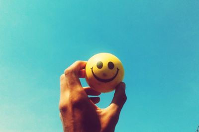 Close-up of hand holding yellow smiley face ball against blue sky