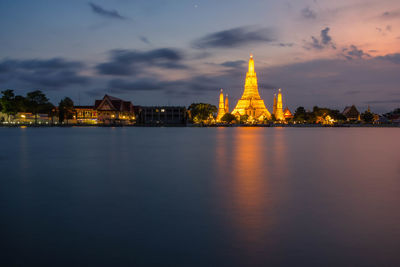 Illuminated buildings at waterfront against cloudy sky