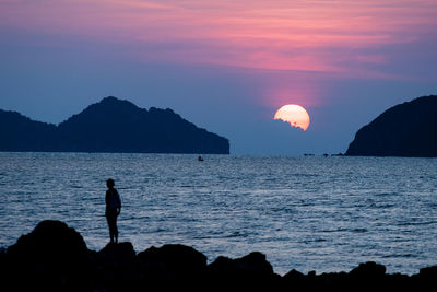 Silhouette person standing on rocks by sea against sky during sunset