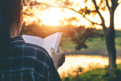 Rear view of man holding book against sun during sunset