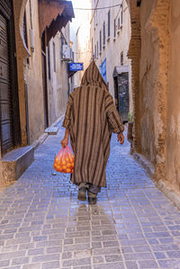 Rear view of man walking in traditional hood clothing while carrying oranges