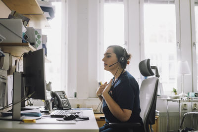 Side view of mature female nurse doing video call while sitting on chair in clinic