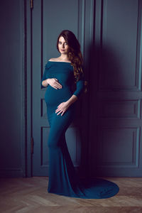 Pregnant woman in a long blue dress standing against the wall in the studio