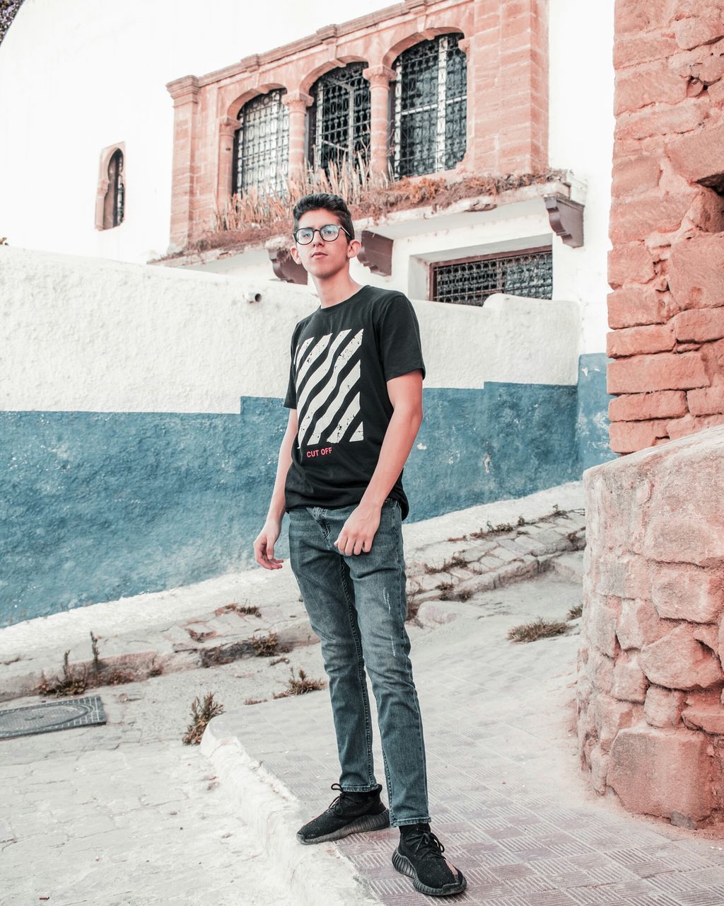 one person, full length, architecture, looking at camera, young adult, portrait, building exterior, standing, real people, casual clothing, front view, built structure, wall, hands in pockets, lifestyles, young men, day, glasses, fashion, outdoors, jeans