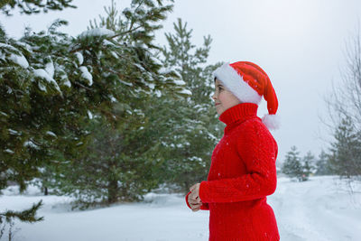 Teenager in a red sweater and a red santa claus hat stands in the winter in the park