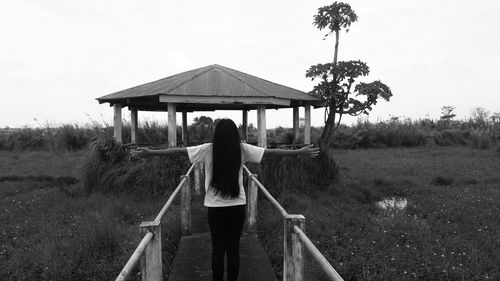 Rear view of woman with arms outstretched amidst field on walkway against gazebo