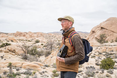 Man with glasses, hat, and camera in desert by stones and plant life