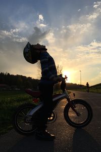 Side view of boy looking up with mountain bike on road against sky during sunset