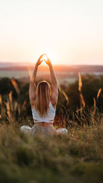 Back view on woman sitting in meditation yoga pose and catching sun by hands at sunset outdoors	
