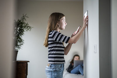 Girl holding mobile phone while using digital tablet on wall with sister sitting in background at smart home