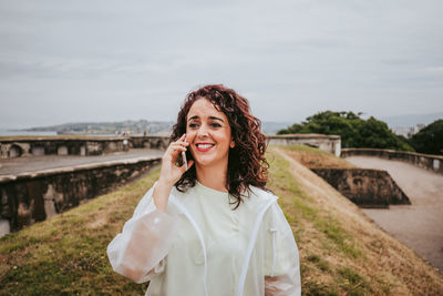 Smiling woman talking over phone while standing on grass against sky