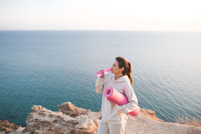 Pretty woman drinking water from pink bottle wear spotswear hoodie and pants hold yoga mat over sea