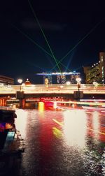 Light trails on river by illuminated city against sky at night