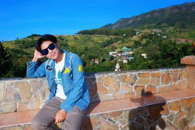 Portrait of man wearing sunglasses while sitting on retaining wall against mountain