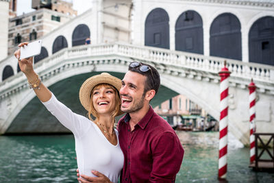 Woman taking selfie with boyfriend against bridge in city