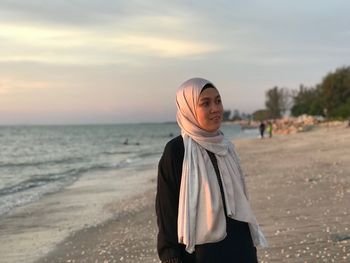 Portrait of young woman standing at beach against sky