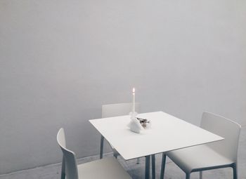 Lit candle on table by empty chairs against wall