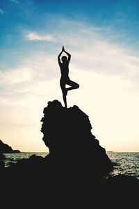 Silhouette woman practicing yoga on rock by sea during sunset