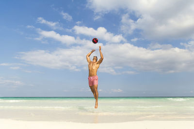 Fit shirtless young man playing with ball while jumping over sea shore against sky