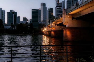 Illuminated buildings by river against sky at dusk