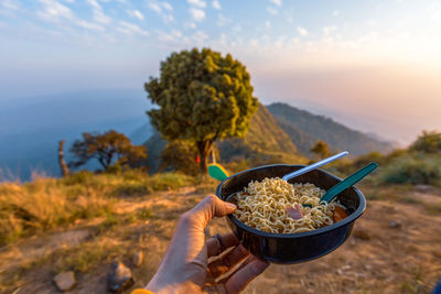 Cropped hand holding bowl with food against mountains during sunset