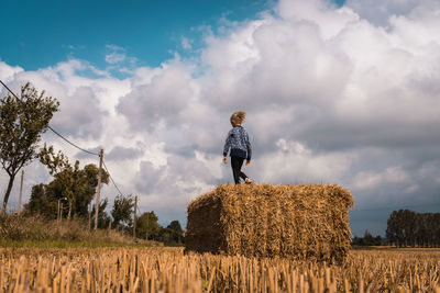 Low angle view of girl walking on hay bale at farm against cloudy sky