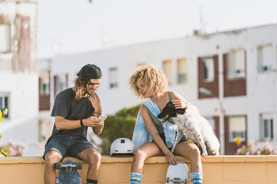 Friends using mobile phone while sitting with dog on retaining wall in city